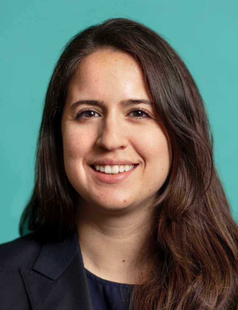 A woman with long brown hair and a dark blazer smiles in front of a teal background.